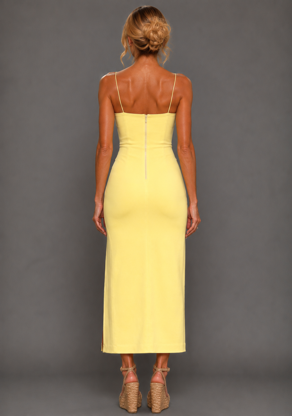 Back image of a model wearing the VSPR Aurora Spaghetti Strap Lemon Midaxi Dress in lemon, revealing the open back, centre-back zip and fine straps, standing against a grey studio backdrop.