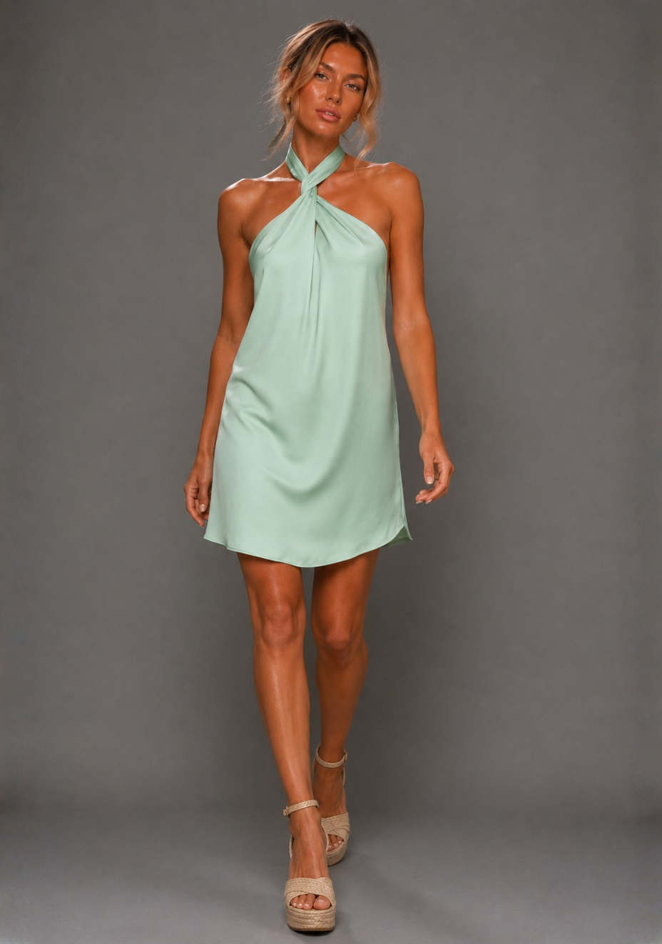 Front image of a model wearing the VSPR Ayesha Twist Neck Sage Mini Dress in Sage Green with a draped twist halterneck and loose, body-skimming silhouette, standing against a grey studio backdrop
