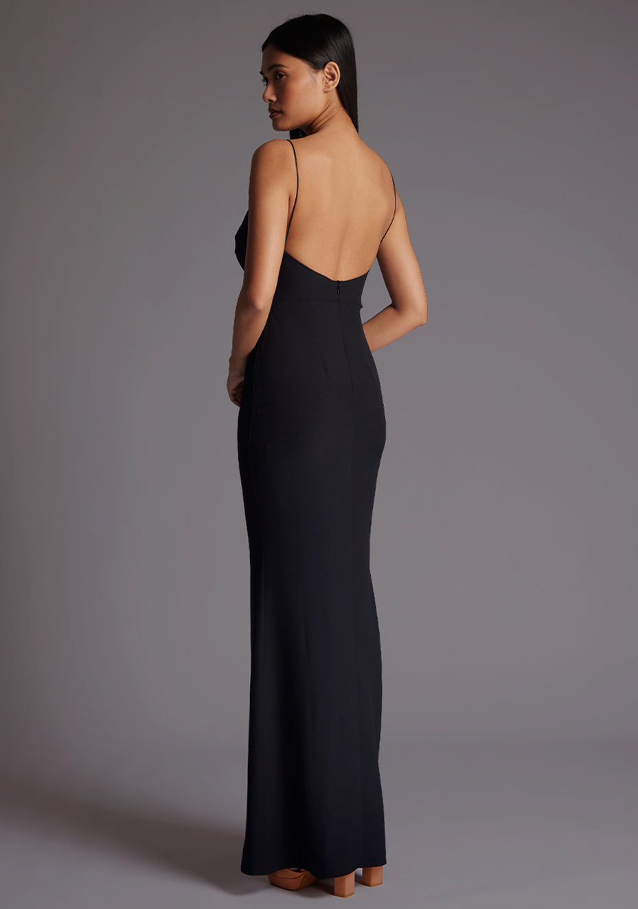 Back quarter image of a model wearing a black maxi dress with a v-neckline, thin straps with twist front detail at bust, low back and subtle flair at hem. The design featured is the Vesper Marlowe Black Maxi Dress