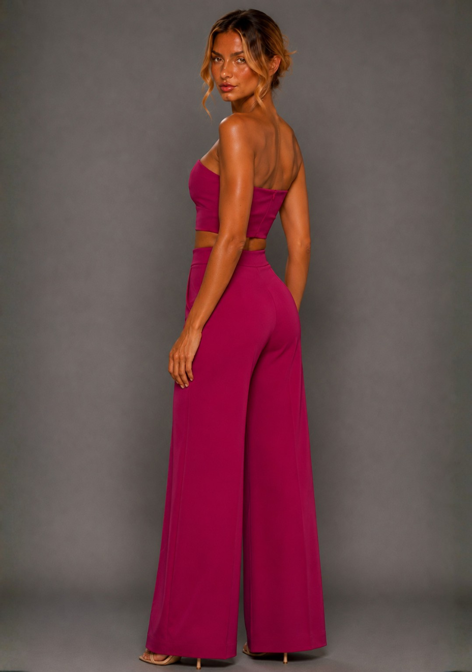 Side back image of a model wearing the VSPR Marty Magenta Wide Leg Trousers, showcasing the high-rise waist and sweeping wide leg, paired with the VSPR Renata Strapless Magenta Crop Top, standing against a grey studio backdrop.