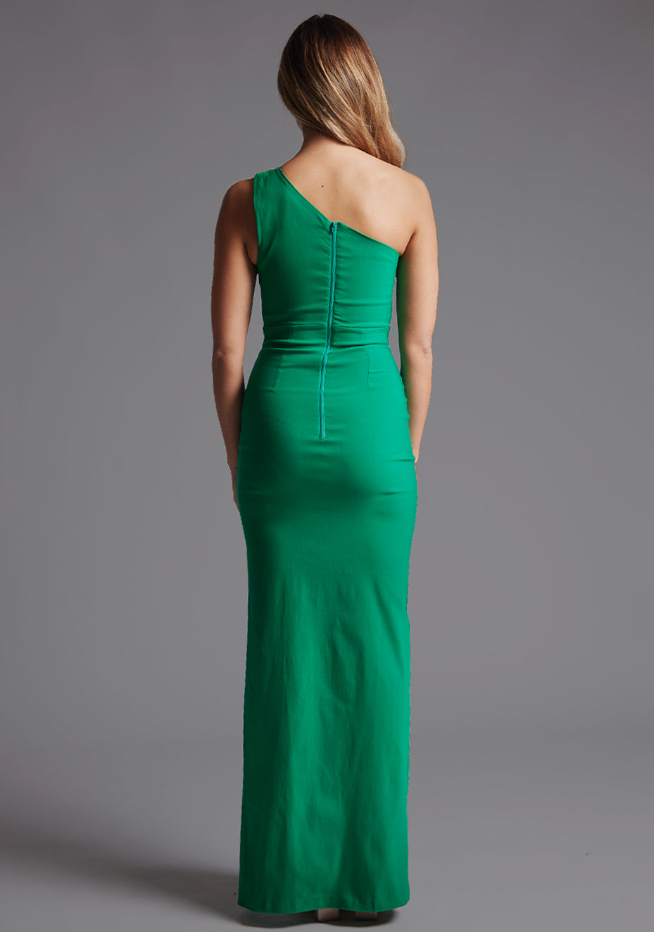 Back image of a model wearing a green one shoulder maxi dress with a one-shoulder design, cut-out detail and centre back zip fastening. The design featured is the Vesper Maya Emerald Green One Shoulder Maxi Dress.