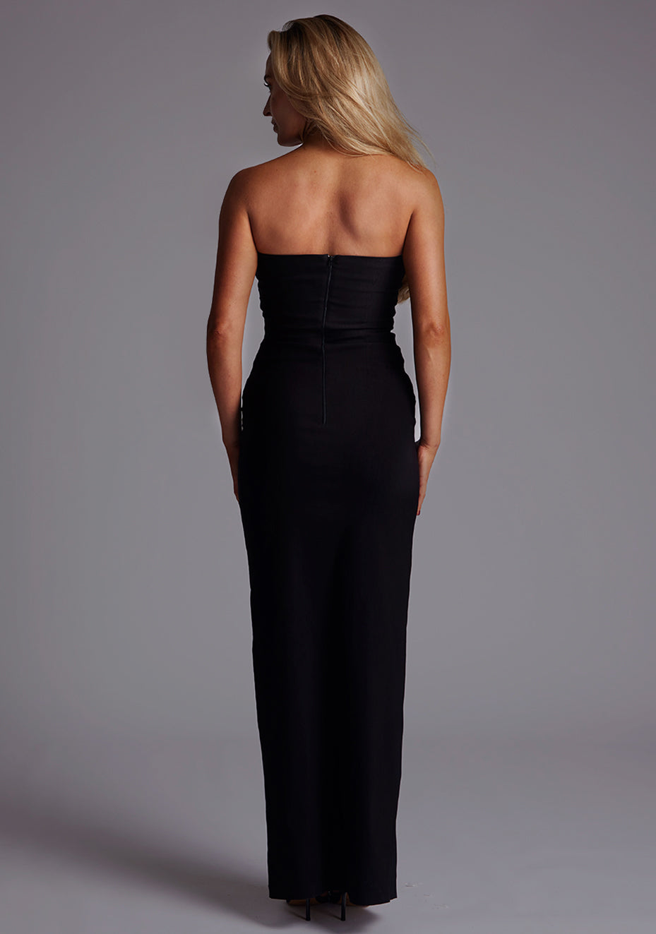 Back image of a model wearing the Vesper Alaya Bandeau Maxi Dress in Black, featuring a clean strapless back, centre-back zip closure, and refined silhouette.