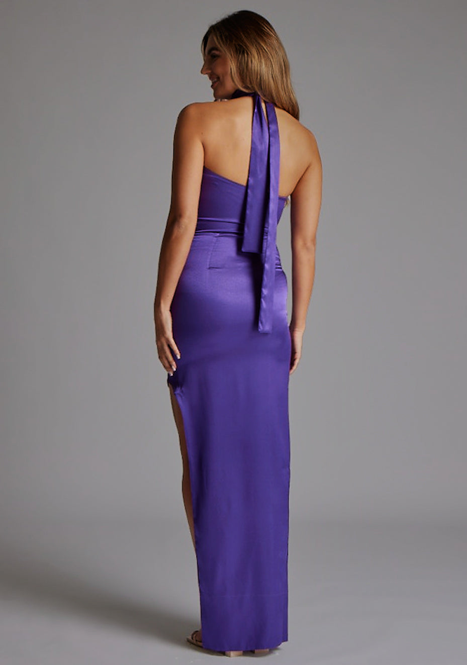 Back image of a model wearing the VSPR Bessie Satin Scarf Detail Purple Maxi Dress in Purple, showcasing the elegant halter straps, defined waist, and flowing maxi silhouette against a grey studio backdrop.