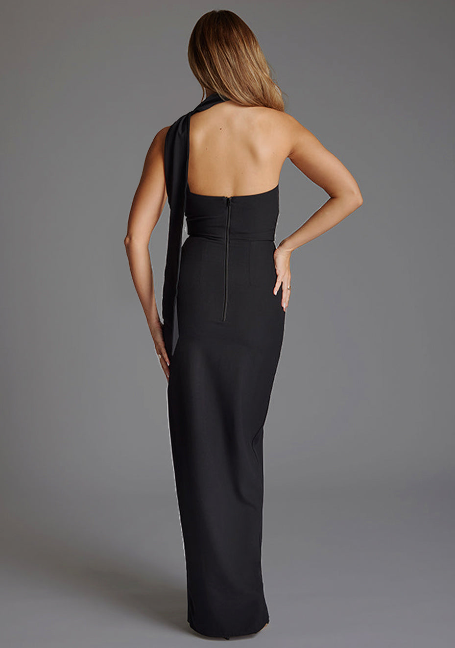 Back image of a model wearing a black maxi evening dress with asymmetric neckline, front skirt split and statement collar drape detail. The design featured is the Vesper Cordula Scarf Detail One Shoulder Maxi Dress Black.