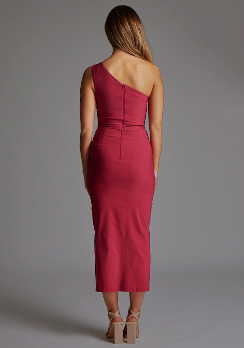 Back image of blonde model wearing a raspberry one shoulder midaxi dress with a asymmetric neckline, one shoulder and front skirt split. The design featured is the Vesper Gianna One Shoulder Midaxi Dress in Raspberry.