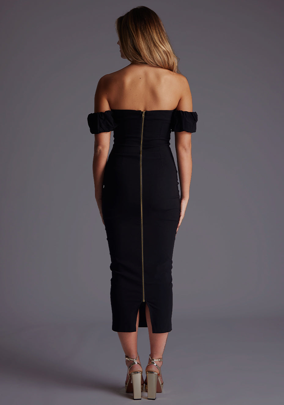 Back image of a model wearing a black bardot midaxi dress with a bardot neckline with off shoulder puff sleeves and a statement gold centre back zip. The design featured is the Vesper Hailey Bardot Neckline Midaxi Dress in Black.