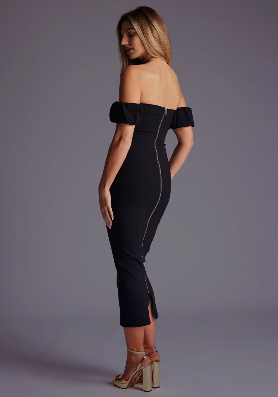 Side back image of a model wearing a black bardot midaxi dress with a bardot neckline with off shoulder puff sleeves and a statement gold centre back zip. The design featured is the Vesper Hailey Bardot Neckline Midaxi Dress in Black.