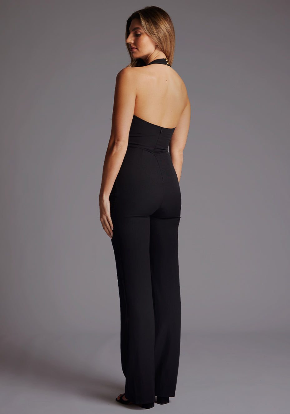 Side back image of a model wearing a black jumpsuit, featuring a deep V-neckline with halter neck style straps, wide leg, and gold buttons at the back of the neck. The dress feature is the Vesper Hazel Halterneck Wide Leg Jumpsuit in Black.