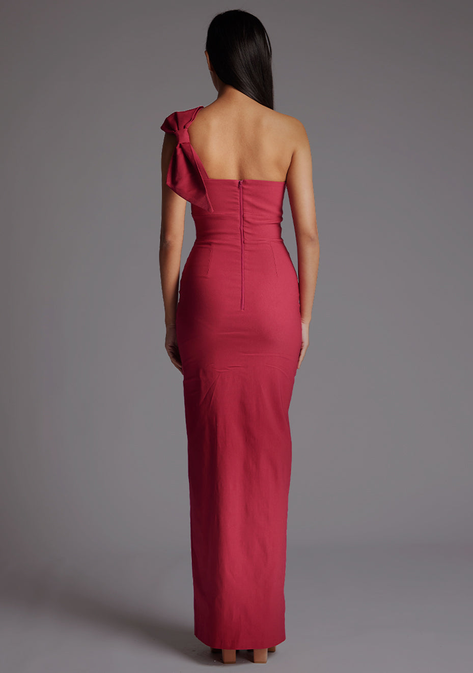 Back image of a model wearing a raspberry maxi dress, featuring an asymmetric neckline, one shoulder with statement bow detail, and a front skirt split. The dress feature is the Vesper Keegan One Shoulder Maxi Dress in Raspberry.