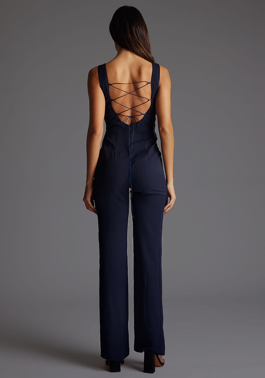Back image of a model wearing a navy jumpsuit wit a high neckline at the front and a open back with strap detailing across the back. The design featured is the Vesper Lilium Lace Up Back Wide Leg Jumpsuit in Navy.