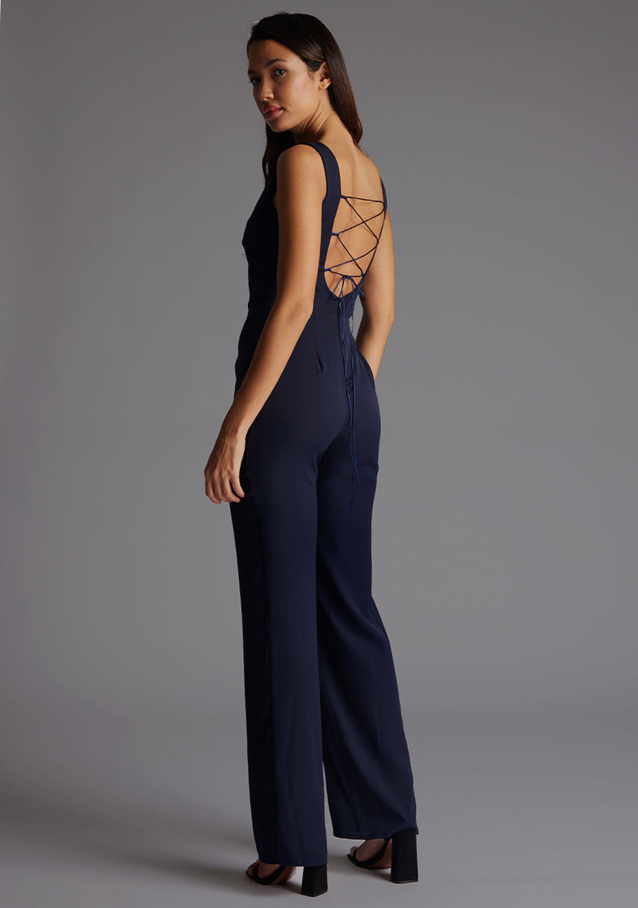 Back quarter image of a model wearing a navy jumpsuit wit a high neckline at the front and a open back with strap detailing across the back. The design featured is the Vesper Lilium Lace Up Back Wide Leg Jumpsuit in Navy.