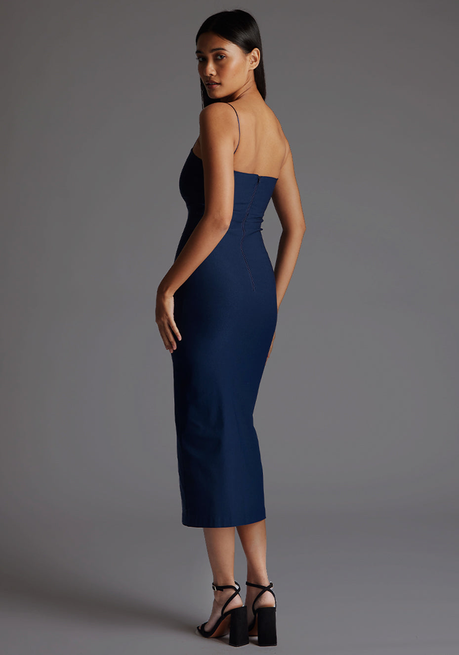 Side back image of model wearing a navy strappy midaxi dress with a sweetheart neckline, thin straps, centre front skirt split and centre back zip for a perfect fit. The design featured is the Vesper Miren Strappy Bodycon Midaxi Dress in Navy.