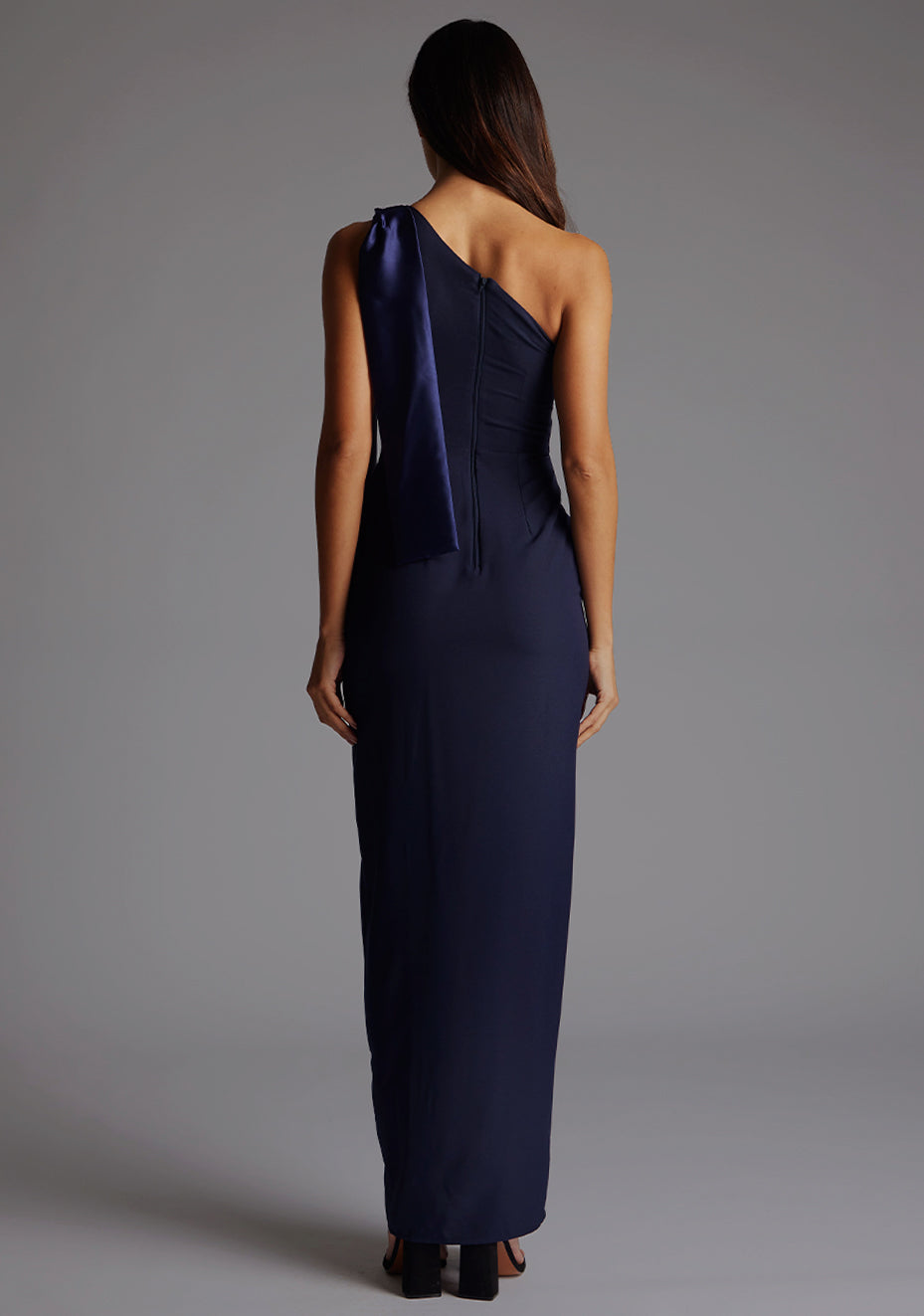 Back image of a model wearing a One Shoulder Navy Maxi Dress with a one shoulder contrast satin band and bow detail with a front skirt split. The design features Vesper Olympia One Shoulder Maxi Dress in Navy.