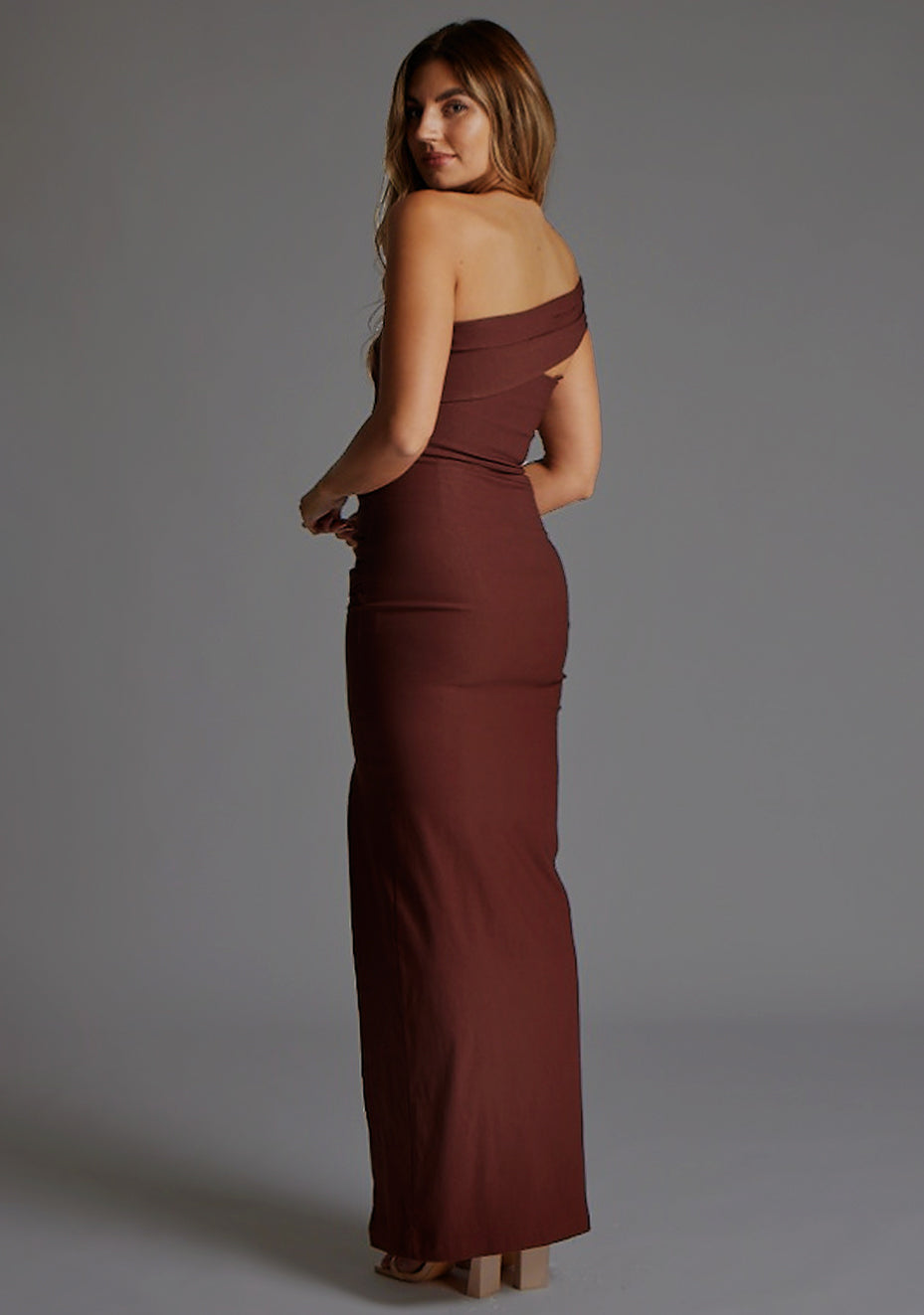 Side back image of a blonde model wearing a Chocolate Maxi Dress with an asymmetric neckline and one-sleeve design and front skirt split adds allure. The design features Vesper Pearson Bardot Ruched Maxi Dress in Chocolate.