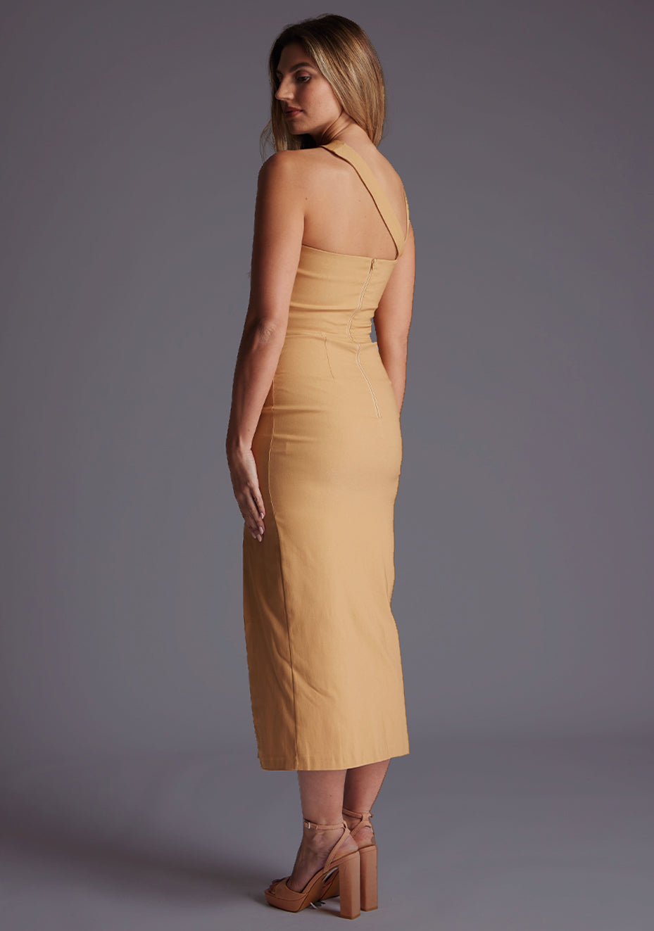 Back quarter image of a blonde model wearing a coffee midaxi dress with a one shoulder design, tasteful cutout detailing, and a front skirt split. The design featured is the Vesper Cerellia Iced Coffee Midaxi Dress