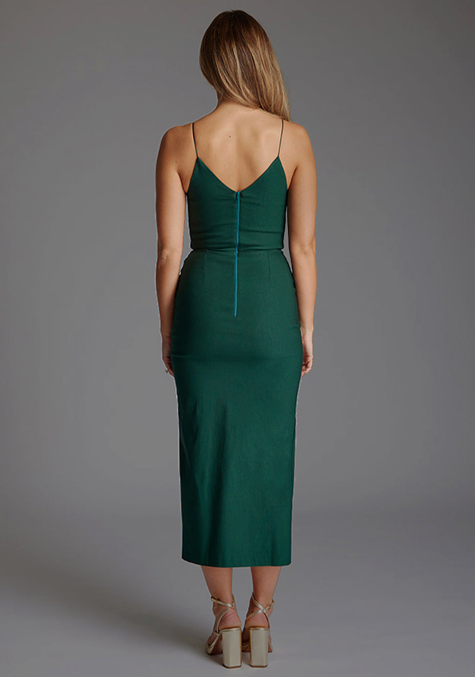 Back image of blonde model wearing a forest green midaxi dress with a plunge neckline, delicate thin straps for a feminine touch, and front split that adds a modern edge. The design featured is the Vesper Palmer Forest Green Midaxi Dress