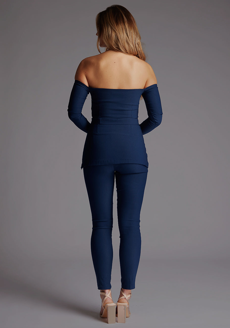 Back image of a model wearing a navy top paired with matching navy trousers. The top features a bardot neckline with soft sweetheart effect, long sleeves and a belt.The styles featured are Vesper Parisa Navy Long Sleeved Bardot Top and Vesper Danica Navy Trousers