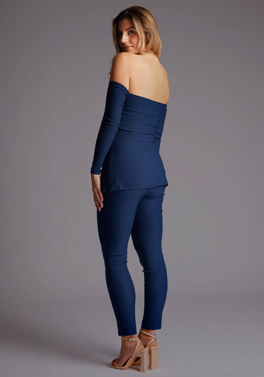 Back quarter image of a model wearing a navy top paired with matching navy trousers. The top features a bardot neckline with soft sweetheart effect, long sleeves and a belt.The styles featured are Vesper Parisa Navy Long Sleeved Bardot Top and Vesper Danica Navy Trousers