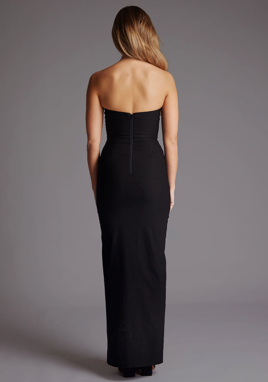 Back image of a blonde model wearing a black bodycon maxi dress with a strapless elegance with a front skirt split for graceful movement and flattering slim fit enhances silhouette. The design featured is the Vesper Remington Black Bodycon Maxi Dress.