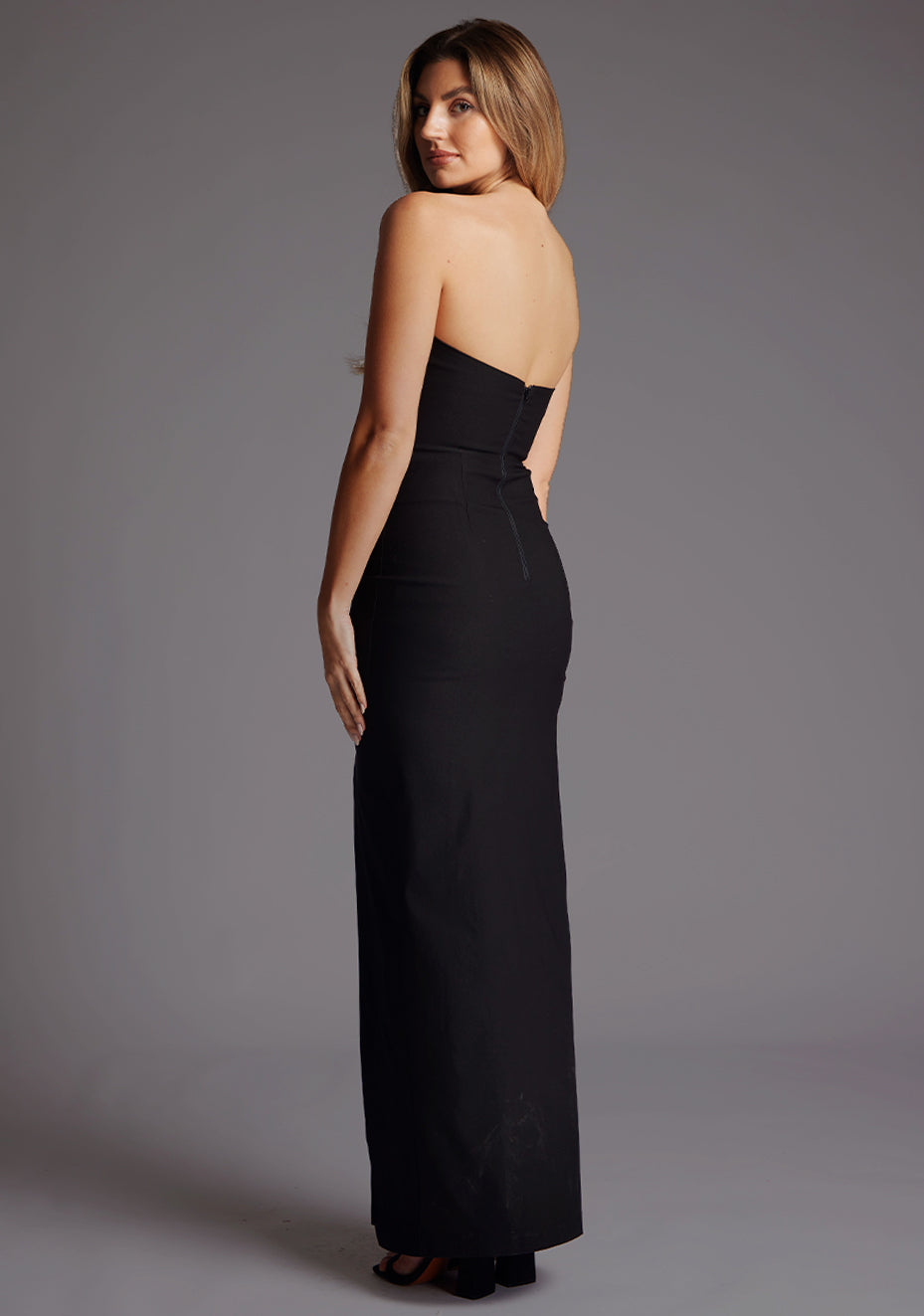 Back quarter image of a blonde model wearing a black bodycon maxi dress with a strapless elegance with a front skirt split for graceful movement and flattering slim fit enhances silhouette. The design featured is the Vesper Remington Black Bodycon Maxi Dress.