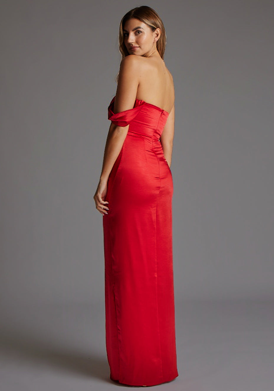 Back quarter image of a blonde model wearing a Red Maxi Dress with a one-sleeve off-shoulder design, crafted with luxurious satin fabric, a design features Vesper Roselie Red Maxi Dress