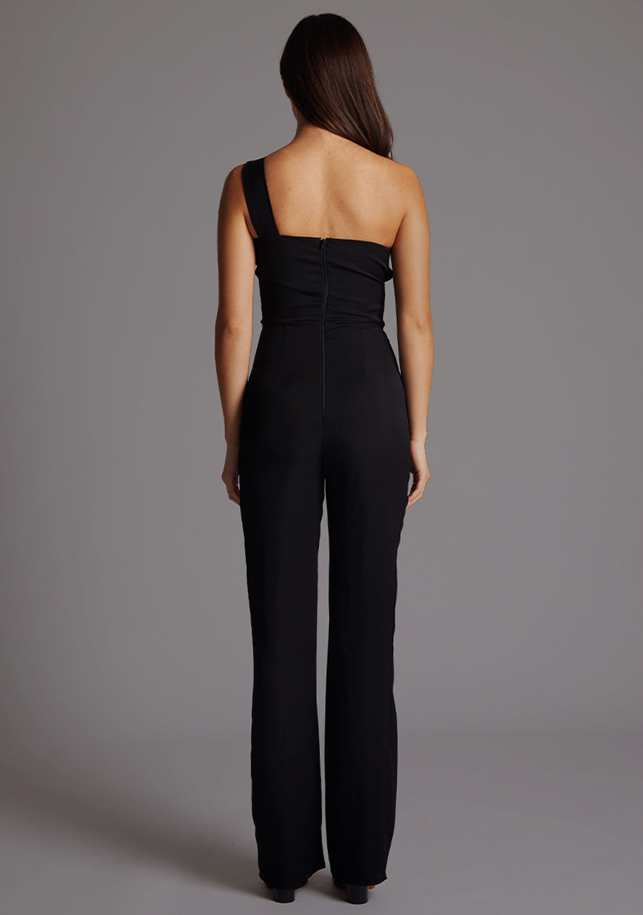 Back image of a model wearing a black Jumpsuit with a stylish one-shoulder design, accentuated by a sophisticated front cut-out, slim fit and convenient back zip. The design featured is the Vesper Rosemary Black Jumpsuit.