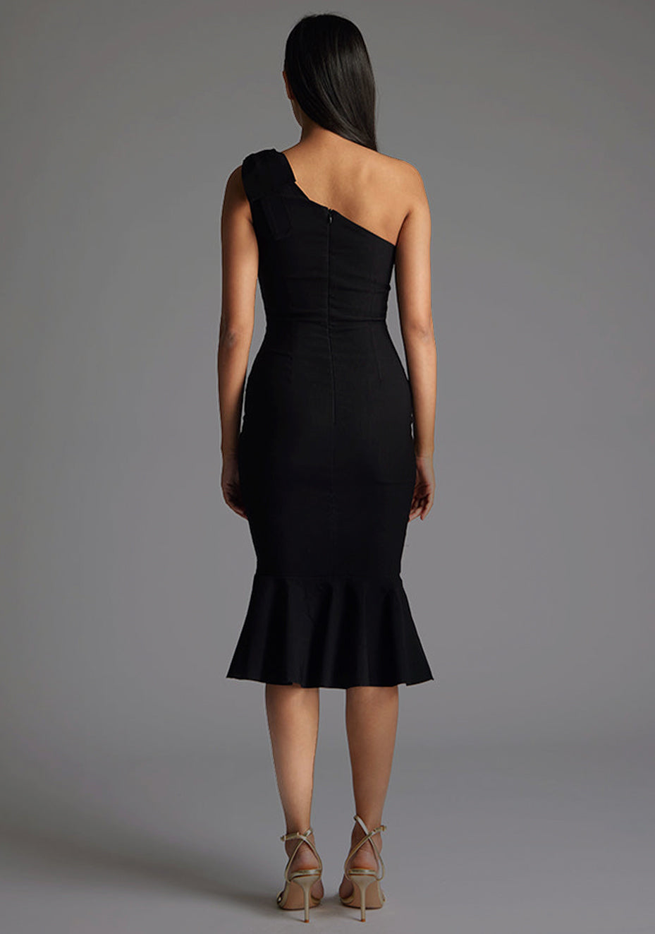 Back image of a model wearing a black one shoulder midaxi dress with a asymmetric neckline, playful one-shoulder with bow detail, and frill hem. The design featured is the Vesper Savannah Black One Shoulder Midaxi Dress.