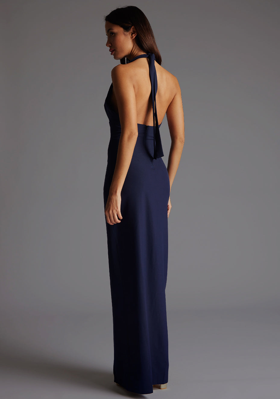 Back quarter image of a blonde model wearing a navy maxidress with a v-plunging neckline, halter design and a front skirt split. The design featured is the Vesper Sedona Navy Maxi Dress.