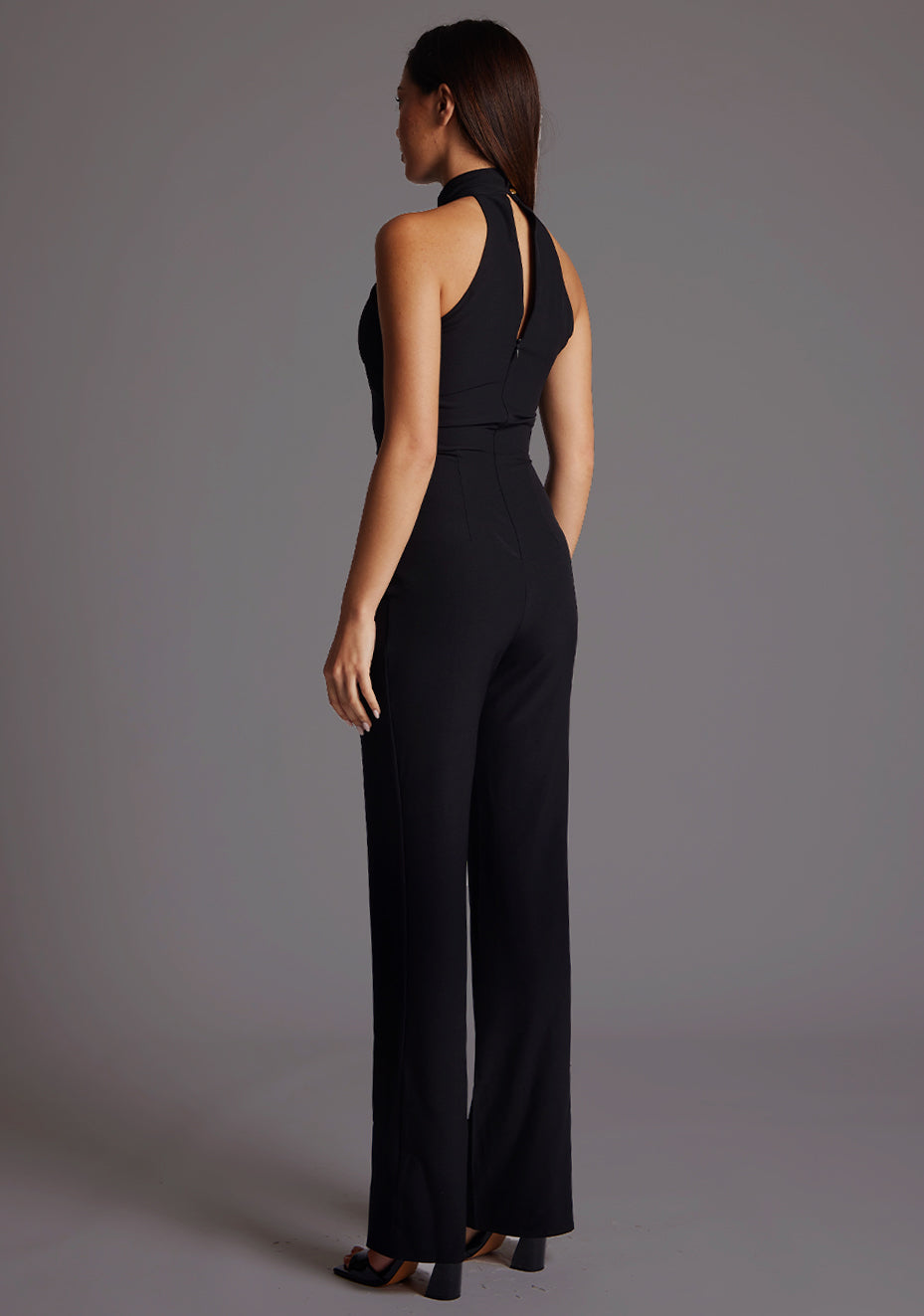 Back quarter image of a model wearing a black jumpsuit with a high neckline, with a v-shaped cut-out at front bust, wide leg and invisible centre back zip. The design featured is the Vesper Stephanie Black Wide Leg Jumpsuit.