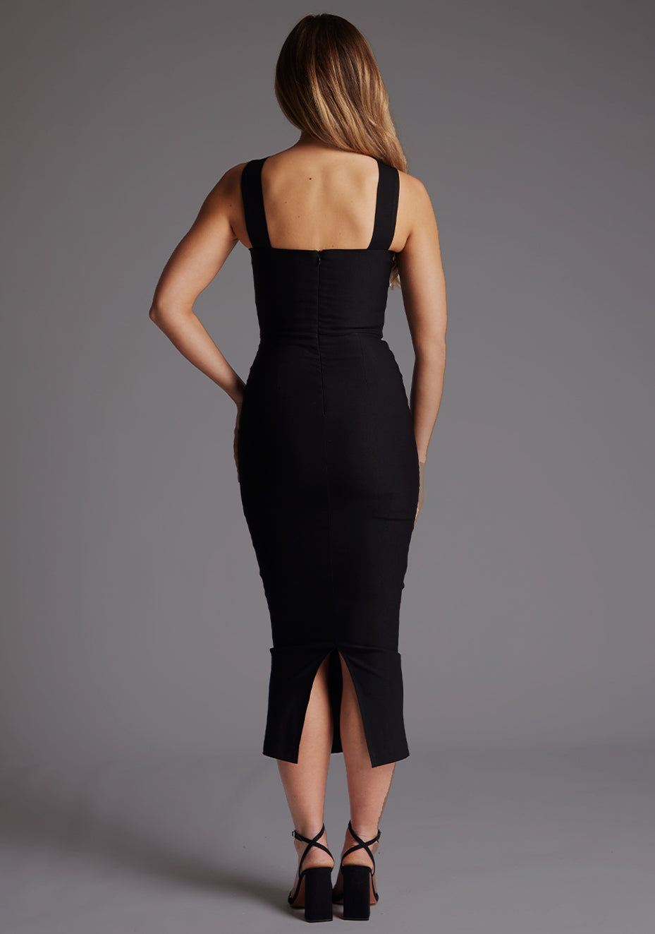 Back image of a model wearing a black midaxi dress with a bodycon fit, sweetheart neckline with wide straps from centre of bust to over shoulder. The design featured is the Vesper Suzanne Black Midaxi Dress.