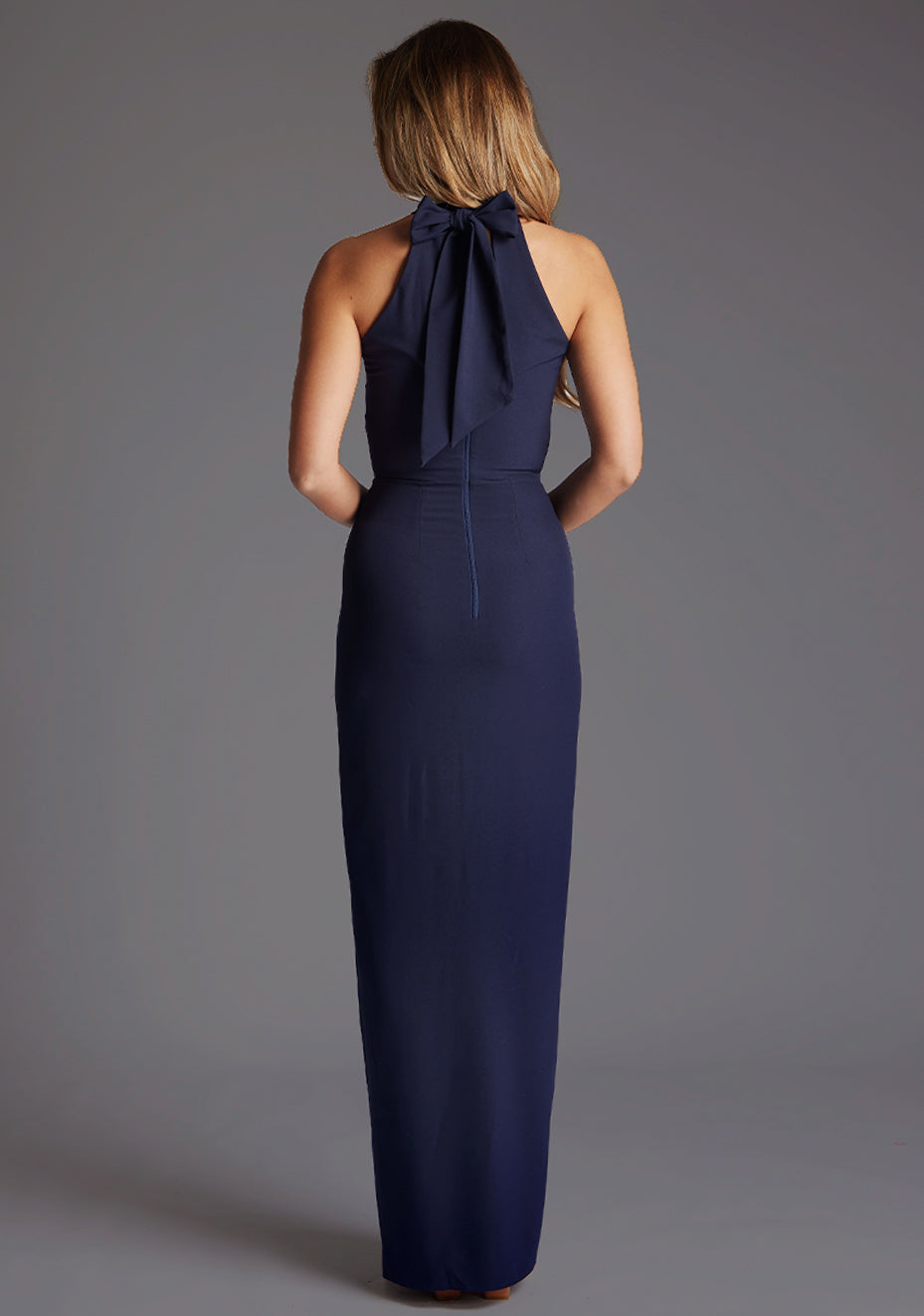Back image of a model wearing a navy maxi dress with a halter neck design, front skirt split for added drama and back zip for easy wear. The design featured is the Vesper Sydney Navy Maxi Dress.