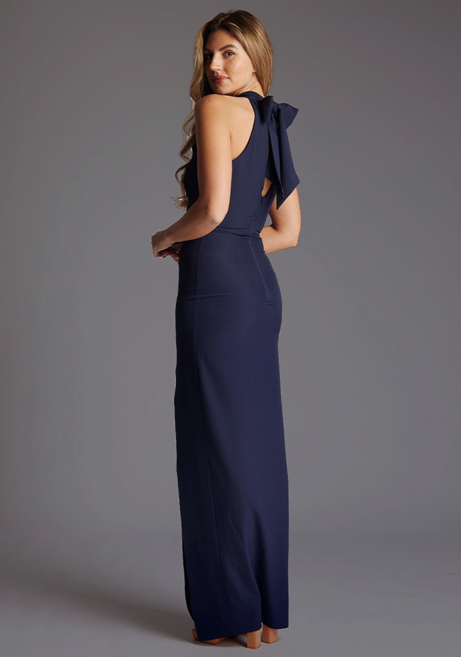 Back quarter image of a model wearing a navy maxi dress with a halter neck design, front skirt split for added drama and back zip for easy wear. The design featured is the Vesper Sydney Navy Maxi Dress.