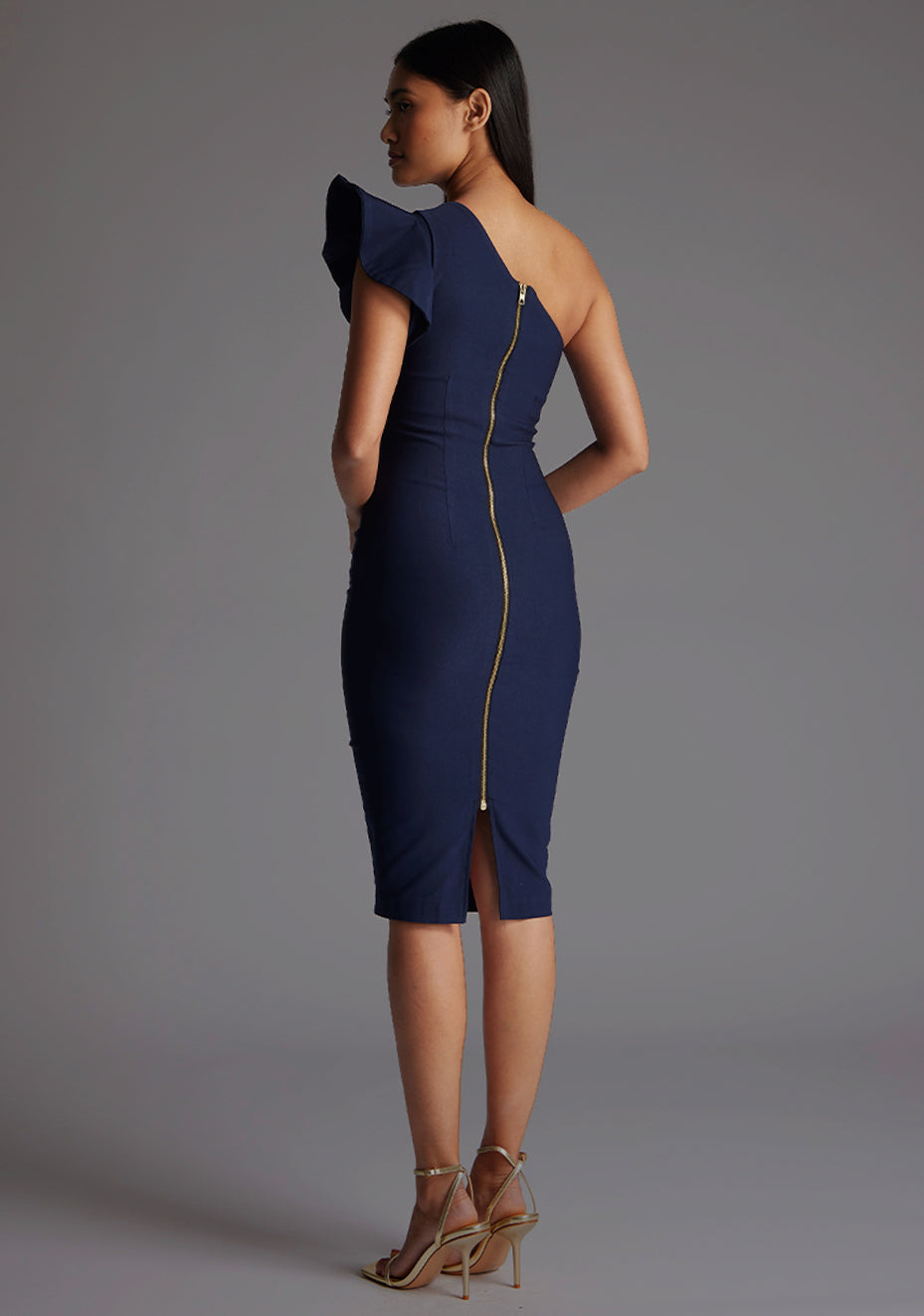 Back quarter image of a model wearing a navy one shoulder midi dress with a stunning one-shoulder design with ruffle detail and an asymmetric neckline, all tied together with a gold centre back zip. The design featured is the Vesper Zanab Navy One Shoulder Midi Dress.