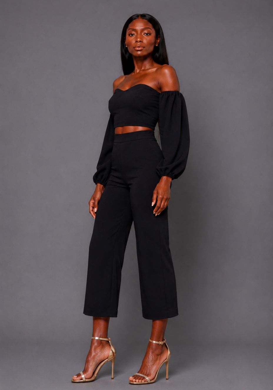 Side back image of a model wearing the VSPR Mindi Wide Leg Black Cropped Trousers, showcasing the smooth high-rise fit and flowing wide-leg shape against a grey studio backdrop.