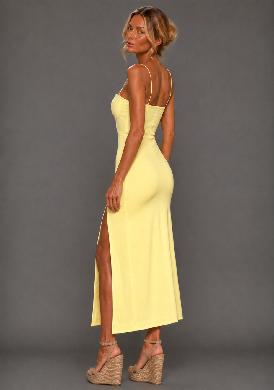 Side back image of a model wearing the VSPR Aurora Spaghetti Strap Lemon Midaxi Dress in lemon, showcasing the fitted silhouette, slender straps and midaxi length, standing against a grey studio backdrop.