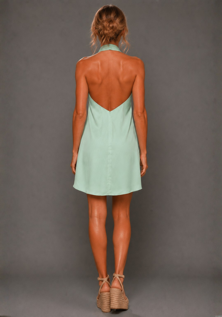 Back image of a model wearing the VSPR Ayesha Twist Neck Sage Mini Dress in Sage Green, highlighting the low V-back finish and relaxed, body-skimming fit, standing against a grey studio backdrop