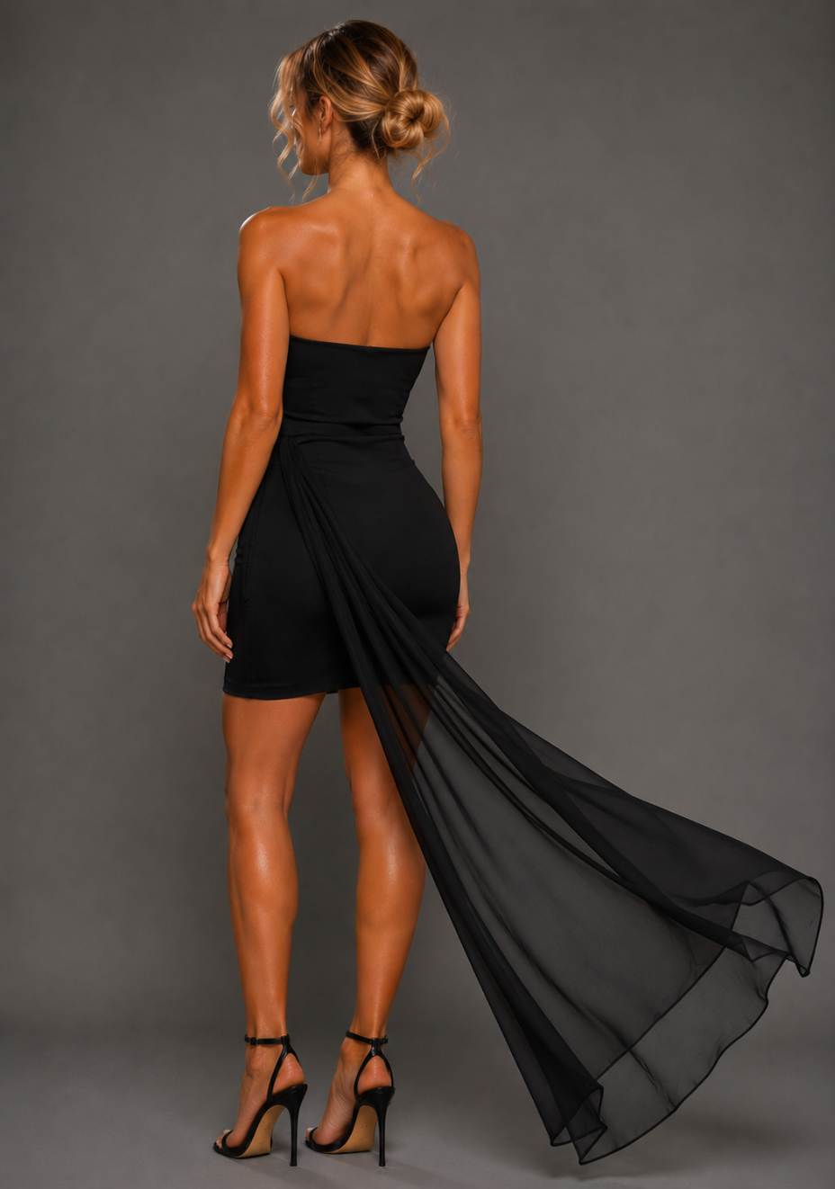 Side back image of a model wearing the VSPR Belisse Strapless Black Mini Dress in Black, showcasing the sleek fitted shape and flowing drape detail from the side against a grey studio backdrop.