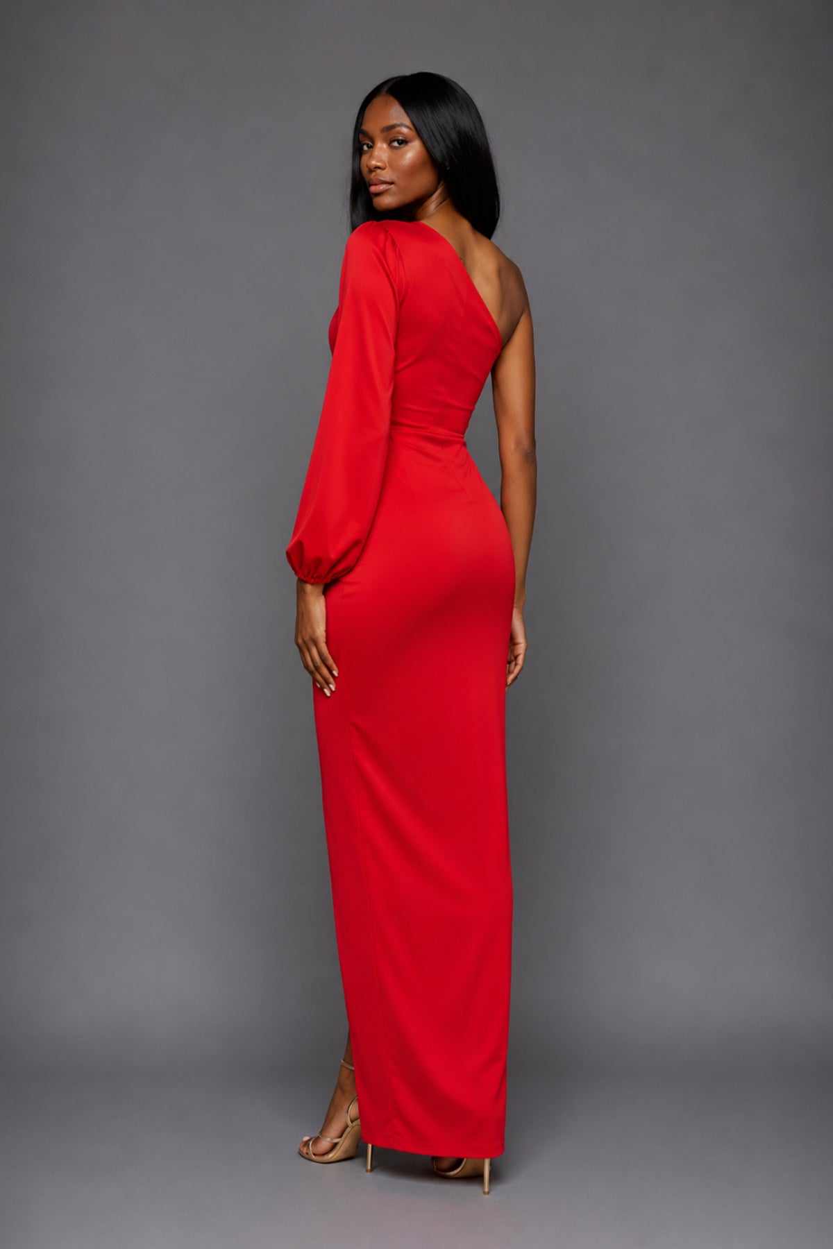 Side-back image of a model wearing the VSPR Keaton One Sleeve Red Maxi Dress, highlighting the sleek silhouette and flowing maxi length from a side angle against a grey studio backdrop.