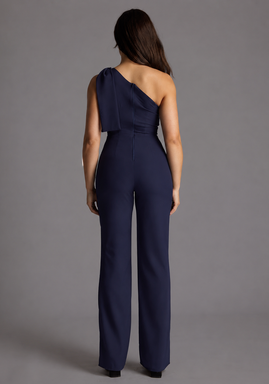 Woman wearing the Vesper Petite Gracie navy one-shoulder jumpsuit against a grey background, back view