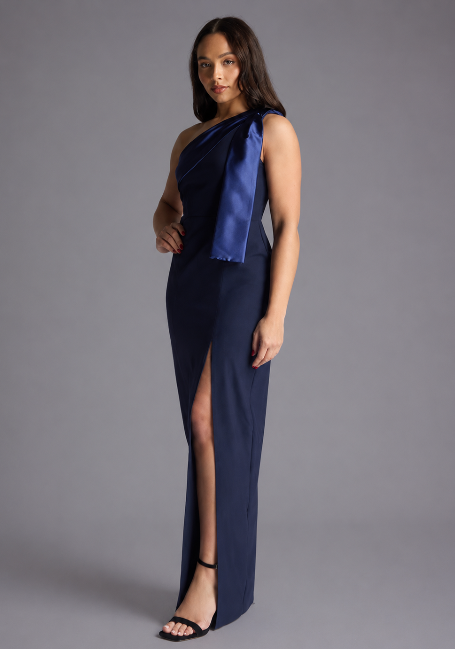 Woman wearing the Vesper Petite Olympia navy one shoulder satin contrast maxi dress against a grey background, side front view