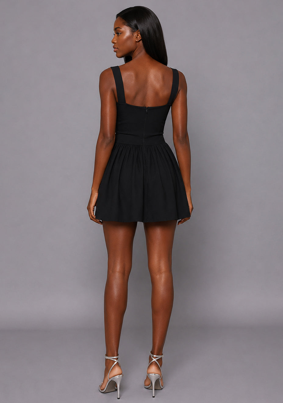 Back image of a model wearing the VSPR Sofia Square Neck Skater Skirt Black Mini Dress showcasing the elegant scoop back design, fitted upper silhouette, and gathered mini skirt against a grey studio backdrop.