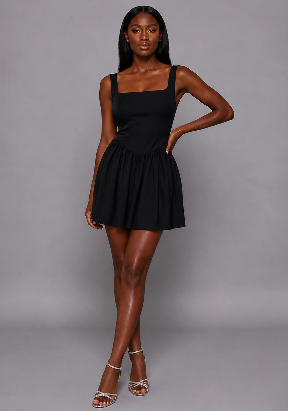 Front image of a model wearing the VSPR Sofia Square Neck Skater Skirt Black Mini Dress featuring a square neckline, fitted bodice, and softly gathered skater skirt, standing against a grey studio backdrop.