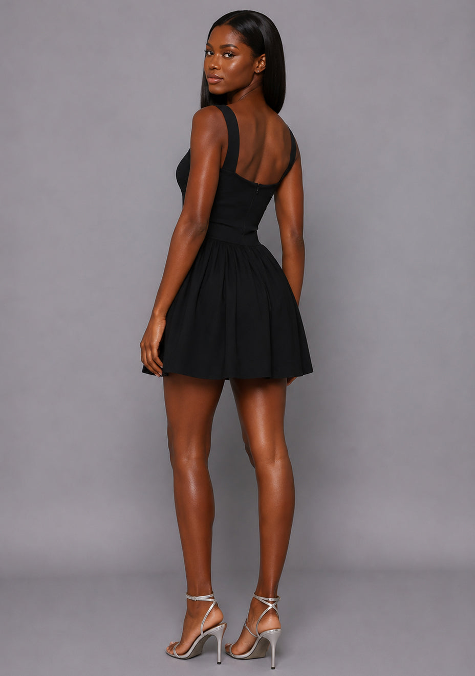 Side back image of a model wearing the VSPR Sofia Square Neck Skater Skirt Black Mini Dress featuring an open scoop back, wide straps, and flowing skater skirt against a grey studio backdrop.