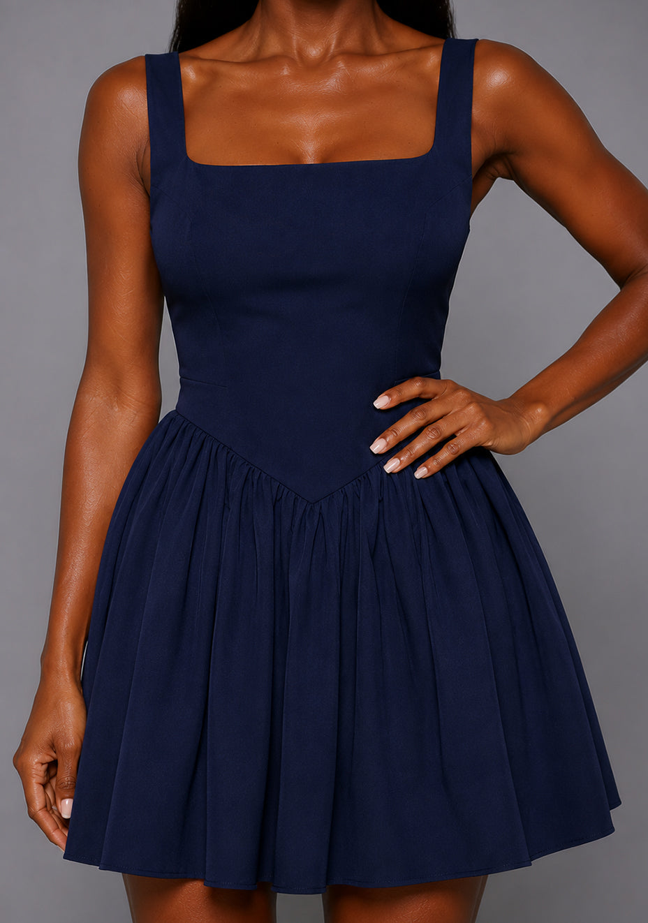 Close up image of a model wearing the VSPR Sofia Square Neck Skater Skirt Navy Mini Dress featuring a structured square neckline, wide shoulder straps, fitted bodice, and gathered skater skirt, standing against a grey studio backdrop.