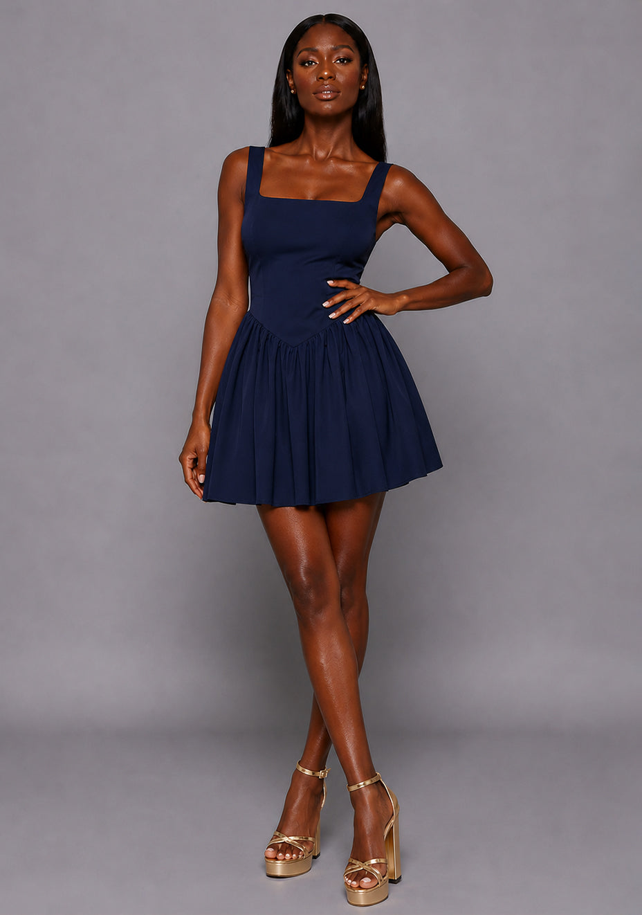 Front image of a model wearing the VSPR Sofia Square Neck Skater Skirt Navy Mini Dress featuring a structured square neckline, wide shoulder straps, fitted bodice, and gathered skater skirt, standing against a grey studio backdrop.