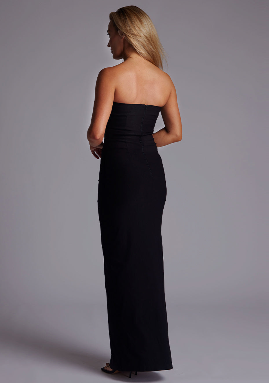Side back image of a model wearing the Vesper Alaya Bandeau Maxi Dress in Black, revealing the smooth strapless back, tailored fit, and centre-back zip.