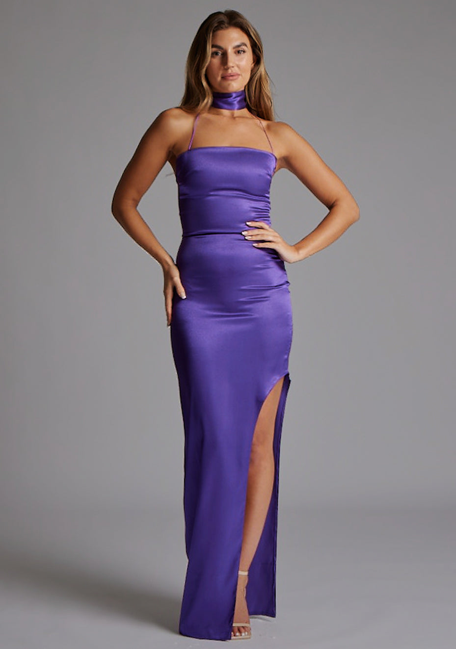 Front image of a model wearing the VSPR Bessie Satin Scarf Detail Purple Maxi Dress in Purple with a sleek straight neckline, halter straps, defined waist, and sweeping maxi length, standing against a grey studio backdrop.