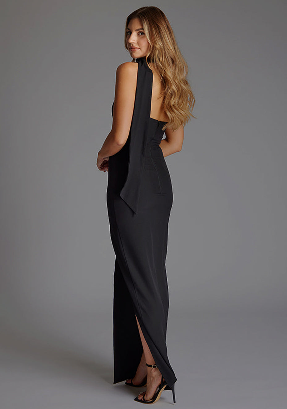 Side back image of a model wearing a black maxi evening dress with asymmetric neckline, front skirt split and statement collar drape detail. The design featured is the Vesper Cordula Scarf Detail One Shoulder Maxi Dress  Black.