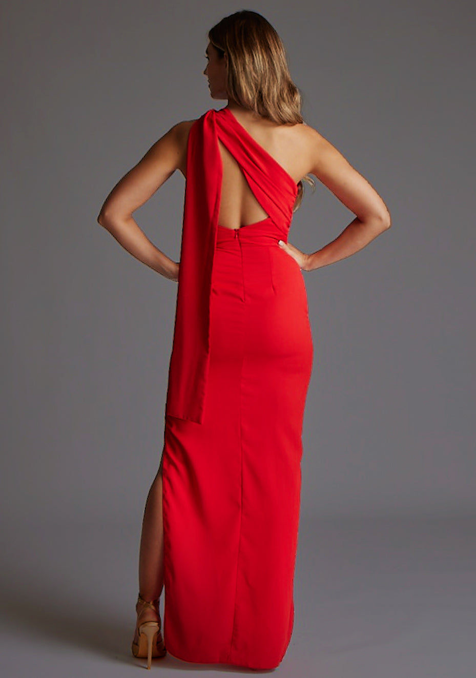 Back image of a model wearing a red maxi dress with a one shoulder design with draped fabric at shoulder and statement asymmetric back with cut-out details and front skirt split.  The design featured is the Vesper Drew One Shoulder Drape Detail Maxi Dress in Red.