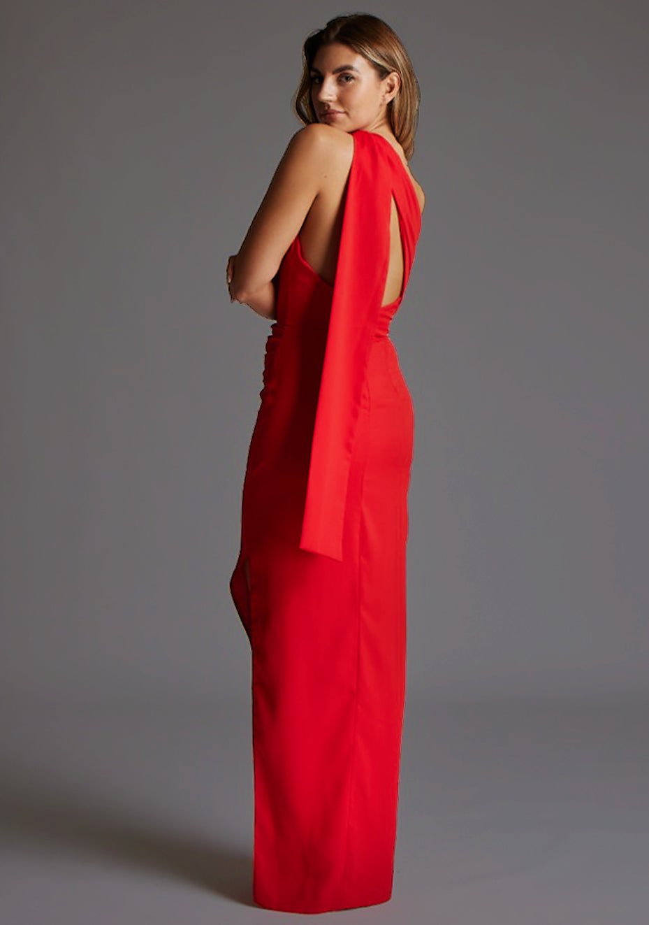 Side back image of a model wearing a red maxi dress with a one shoulder design with draped fabric at shoulder and statement asymmetric back with cut-out details and front skirt split.  The design featured is the Vesper Drew One Shoulder Drape Detail Maxi Dress in Red.